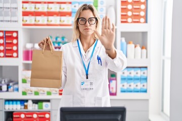 Poster - Young blonde woman working at pharmacy drugstore holding paper bag with open hand doing stop sign with serious and confident expression, defense gesture