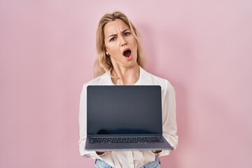 Sticker - Young caucasian woman holding laptop showing screen in shock face, looking skeptical and sarcastic, surprised with open mouth