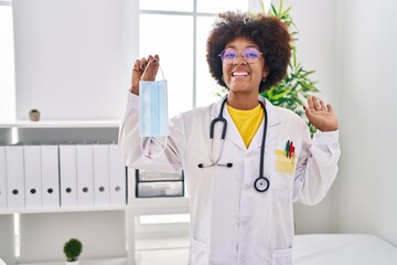 Wall Mural - Young african american woman wearing doctor uniform holding safety mask celebrating achievement with happy smile and winner expression with raised hand