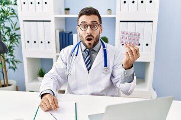 Wall Mural - Handsome hispanic man wearing doctor uniform holding prescription pills scared and amazed with open mouth for surprise, disbelief face