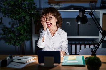 Poster - Middle age woman working at the office at night covering one eye with hand, confident smile on face and surprise emotion.