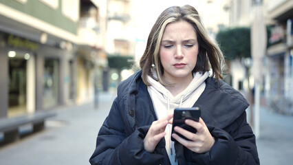 Wall Mural - Young blonde woman using smartphone with serious expression at street