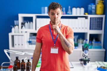 Wall Mural - Young hispanic man working at scientist laboratory asking to be quiet with finger on lips. silence and secret concept.