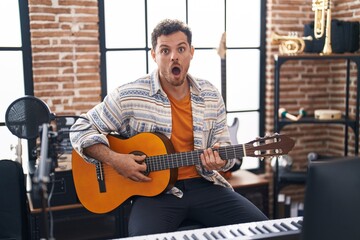Wall Mural - Young hispanic man playing classic guitar at music studio scared and amazed with open mouth for surprise, disbelief face