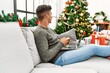 © Krakenimages.com - Young hispanic man using laptop and smartphone sitting on sofa by christmas tree at home