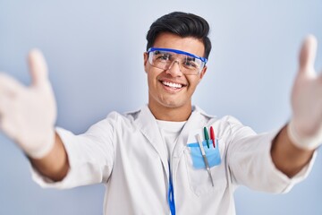 Sticker - Hispanic man working as scientist looking at the camera smiling with open arms for hug. cheerful expression embracing happiness.