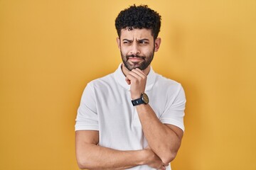 Wall Mural - Arab man standing over yellow background with hand on chin thinking about question, pensive expression. smiling and thoughtful face. doubt concept.