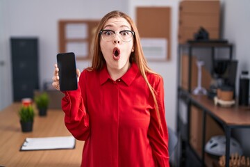 Wall Mural - Redhead woman working at the office showing smartphone screen scared and amazed with open mouth for surprise, disbelief face