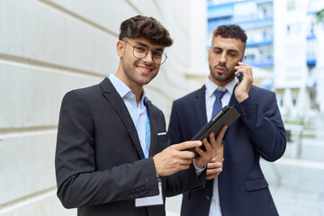 Wall Mural - Two hispanic men business workers using touchpad talking on smartphone at street