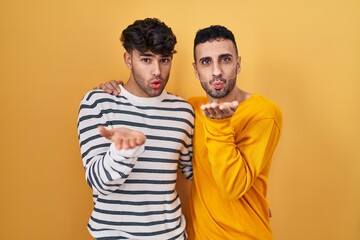 Poster - Young hispanic gay couple standing over yellow background looking at the camera blowing a kiss with hand on air being lovely and sexy. love expression.