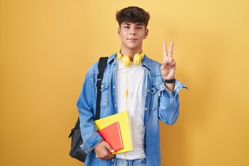 Canvas Print - Hispanic teenager wearing student backpack and holding books smiling with happy face winking at the camera doing victory sign with fingers. number two.