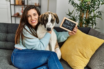 Canvas Print - Young woman holding new home blackboard hugging dog sitting on sofa at home