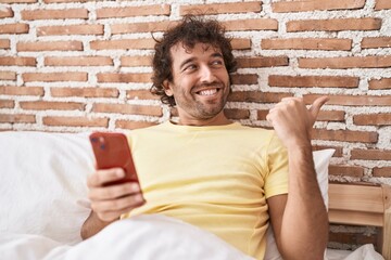 Sticker - Hispanic young man using smartphone on the bed pointing thumb up to the side smiling happy with open mouth