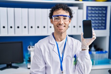 Wall Mural - Hispanic man working at scientist laboratory showing smartphone screen looking positive and happy standing and smiling with a confident smile showing teeth