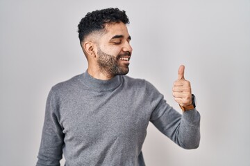 Wall Mural - Hispanic man with beard standing over white background looking proud, smiling doing thumbs up gesture to the side