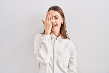 Poster - Young caucasian woman standing over isolated background covering one eye with hand, confident smile on face and surprise emotion.