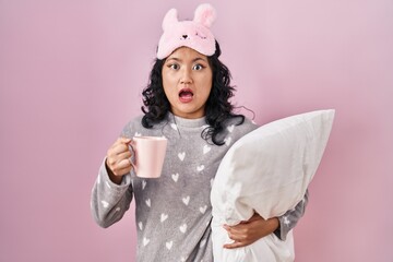 Canvas Print - Young asian woman wearing sleep mask and pajama hugging pillow drinking coffee in shock face, looking skeptical and sarcastic, surprised with open mouth