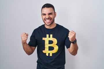 Poster - Young hispanic man wearing bitcoin t shirt very happy and excited doing winner gesture with arms raised, smiling and screaming for success. celebration concept.