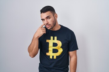 Wall Mural - Young hispanic man wearing bitcoin t shirt pointing to the eye watching you gesture, suspicious expression