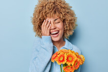 Portrait Of Cheerful Woman With Fair Curly Hair Makes Face Palm Smiles Broadly Holds Orange Gerbera Flowers Dressed In Casual Jumper Isolated Over Blue Background. Spring Time And Womens Day Concept