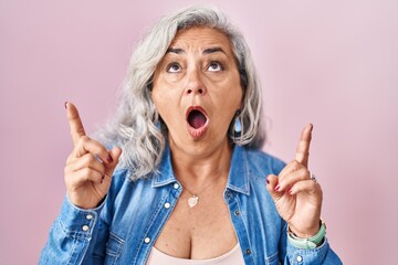 Canvas Print - Middle age woman with grey hair standing over pink background amazed and surprised looking up and pointing with fingers and raised arms.