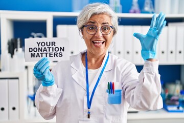 Poster - Middle age woman with grey hair working at scientist laboratory holding your donation matters banner celebrating victory with happy smile and winner expression with raised hands