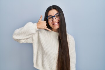 Poster - Young hispanic woman wearing casual sweater over blue background smiling doing phone gesture with hand and fingers like talking on the telephone. communicating concepts.