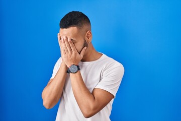 Poster - Young hispanic man standing over blue background with sad expression covering face with hands while crying. depression concept.