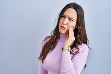 Poster - Young brunette woman standing over blue background touching mouth with hand with painful expression because of toothache or dental illness on teeth. dentist