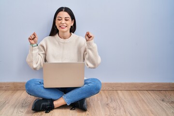 Wall Mural - Young woman using laptop sitting on the floor at home very happy and excited doing winner gesture with arms raised, smiling and screaming for success. celebration concept.