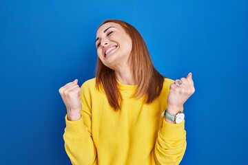 Wall Mural - Young woman standing over blue background celebrating surprised and amazed for success with arms raised and eyes closed. winner concept.