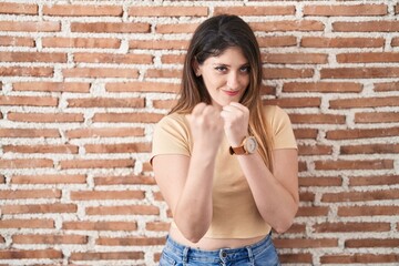 Sticker - Young brunette woman standing over bricks wall ready to fight with fist defense gesture, angry and upset face, afraid of problem