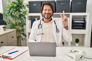 Canvas Print - Handsome middle age doctor man working at the clinic showing and pointing up with fingers number two while smiling confident and happy.