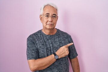 Poster - Middle age man with grey hair standing over pink background pointing with hand finger to the side showing advertisement, serious and calm face