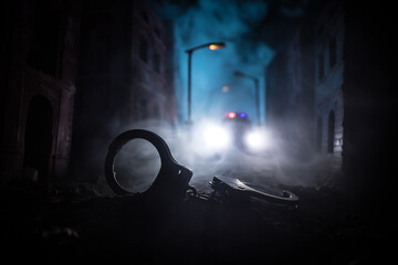 Wall Mural - Police raid at night and you are under arrest concept. Silhouette of handcuffs with police car on backside. Image with the flashing red and blue police lights at foggy background. Slider shot