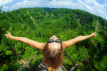 Wall Mural - Family vacation lifestyle. Young woman stand on edge of overhanging balcony on high cliff. Happy girl looking at stunning tropical jungle view. Tukad Melangit is popular travel destination in Bali