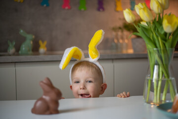 A cute little boy wearing bunny ears on Easter day wants to eat a chocolate easter bunny. A child plays egg hunt for Easter. Charming child celebrates Easter at home.