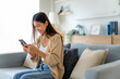 © Monster Ztudio - Happy young asian woman relax on comfortable couch at home texting messaging on smartphone, smiling girl use cellphone chatting, browse wireless internet on gadget, shopping online from home