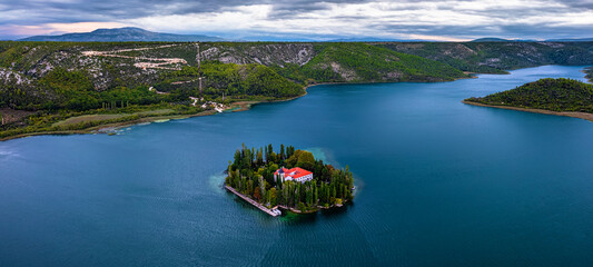 Wall Mural - Visovac, Croatia - Aerial panoramic view of Visovac Christian monastery in Krka National Park on a summer morning with the peaks of Kijevski Kozjak and cloudy sky at background