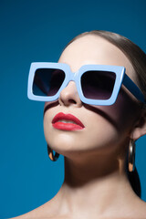 Wall Mural - Fashion and make-up concept. Close-up beautiful woman portrait with classic hairstyle, big retro blue sunglasses, red lipstick and golden round earrings in blue studio background. Model looking up