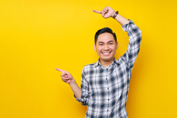 Smiling young handsome Asian man wearing a plaid shirt pointing fingers aside at blank space for advertising text isolated on yellow background. People lifestyle concept