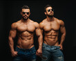 Two shirtless hunks at black background. Fitness models couple in jeans and sunglasses. Two muscular handsome men with six pack abs in studio.