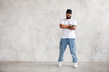 Full length portrait Hispanic man stands arms crossed, in casual wear jeans and white mockup T-shirt and baseball cap, cool guy looks at camera against gray wall