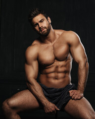 Wall Mural - Brutal sexy man with six pack abs in towel at black background. Handsome fitness male model sitting in studio. Bearded muscled guy covering with towel in studio.