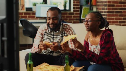 Wall Mural - Happy life partners eating slices of pizza on couch, having fun together watching favorite movie on television. Young man and woman in relationship enjoying delivery food and alcohol.