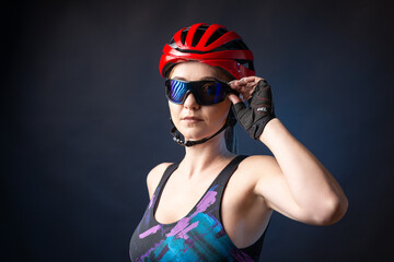 A young female cyclist wearing a safety helmet and glasses, dressed in a bib shorts poses against a black background in the studio.