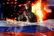 © Arsentyev Vladimir - A soldier in a camouflaged uniform with a rifle against the background of the Flag of Russia and a burning destroyed house, looks into the camera. The concept of war or the game of airsoft