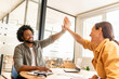 © Vadim Pastuh - Two young happy diverse colleague sitting at desk and taking high five while working together in coworking space, man and woman celebrating successful project completion or good deal