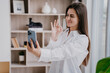 © Iona - Beautiful  brunette girl in white shirt smiling, making selfie at living room, office. Satisfied young woman shows ok sign to boyfriend. Cheerful student holds phone making video call shows ok gesture