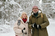 Cheerful senior couple with trekking sticks looking at camera while strolling in winter forest covered with snow on weekend